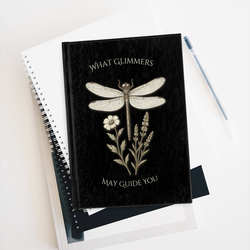 Black notebook with dragonfly and floral design on a white surface. "What Glimmers May Guide You" Dragonfly Journal
