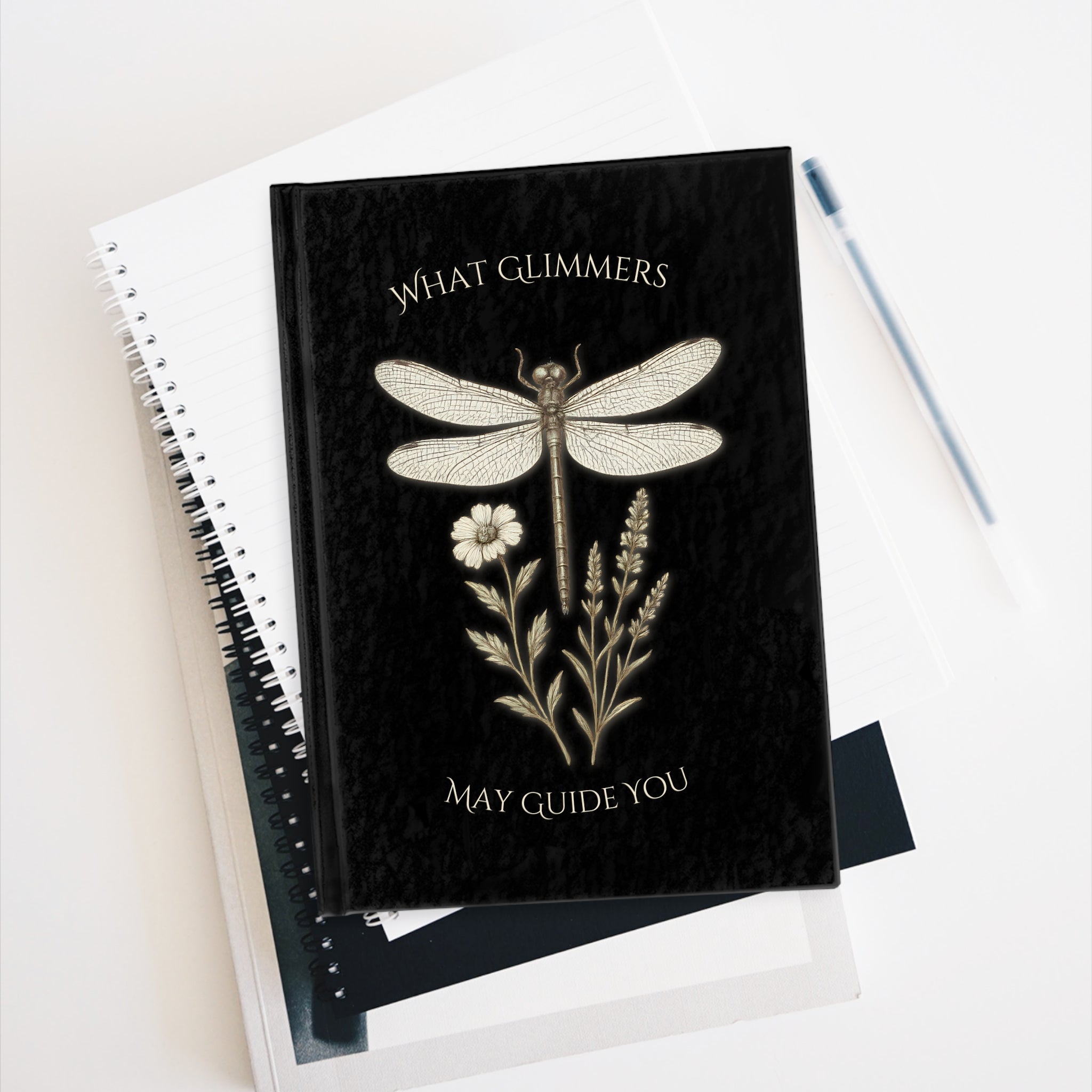 Black notebook with dragonfly and floral design on a white surface. "What Glimmers May Guide You" Dragonfly Journal