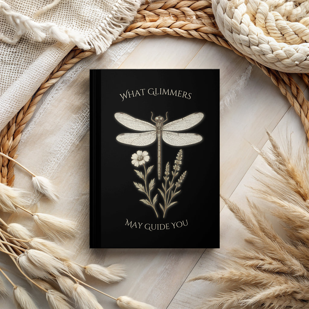 Black notebook with dragonfly design and text on a textured surface with natural elements. "What Glimmers May Guide You" Dragonfly Journal