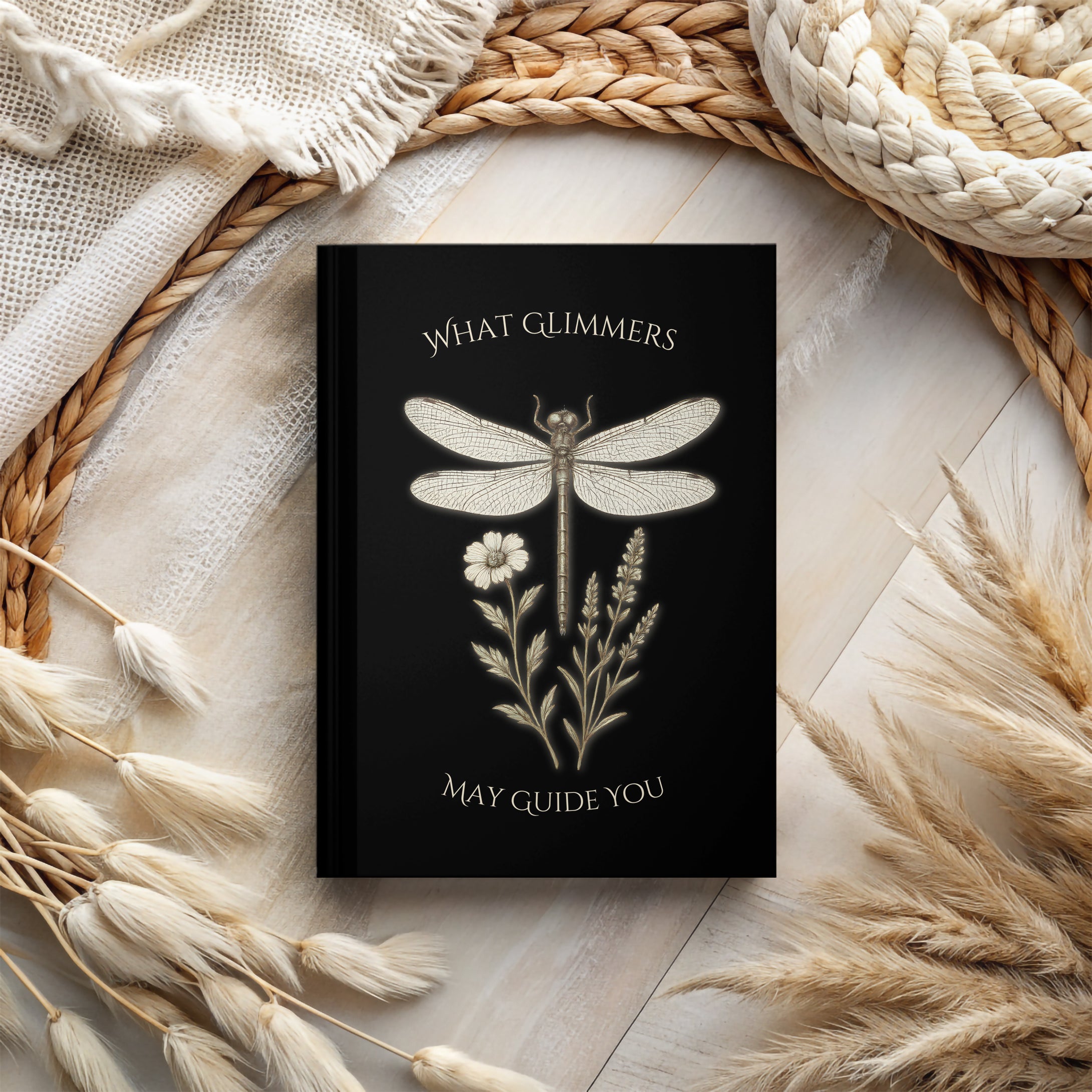 Black notebook with dragonfly design and text on a textured surface with natural elements. "What Glimmers May Guide You" Dragonfly Journal